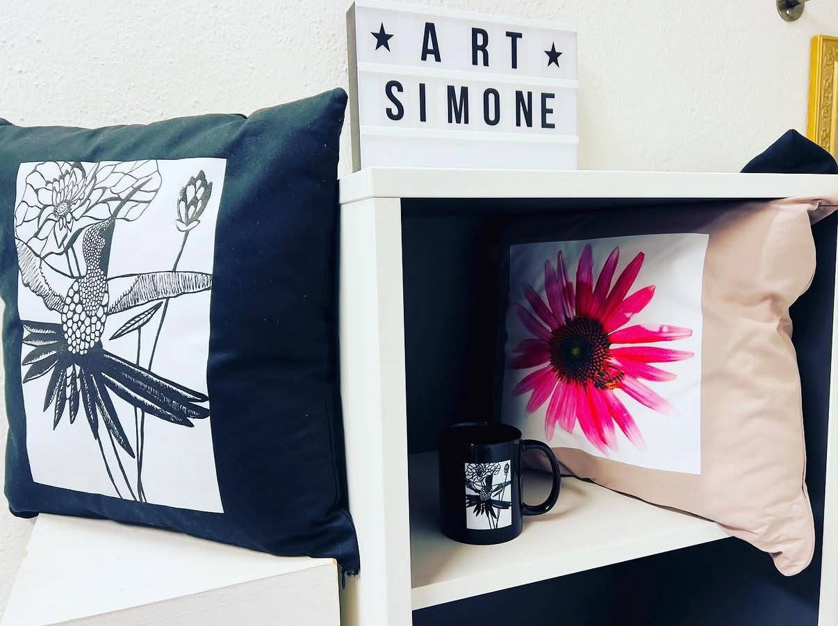 Leuchtschild «ART SIMONE» über einem Regal mit bedruckten Kissen und einer Tasse, alle mit Blüten- und Kranichmotiven
