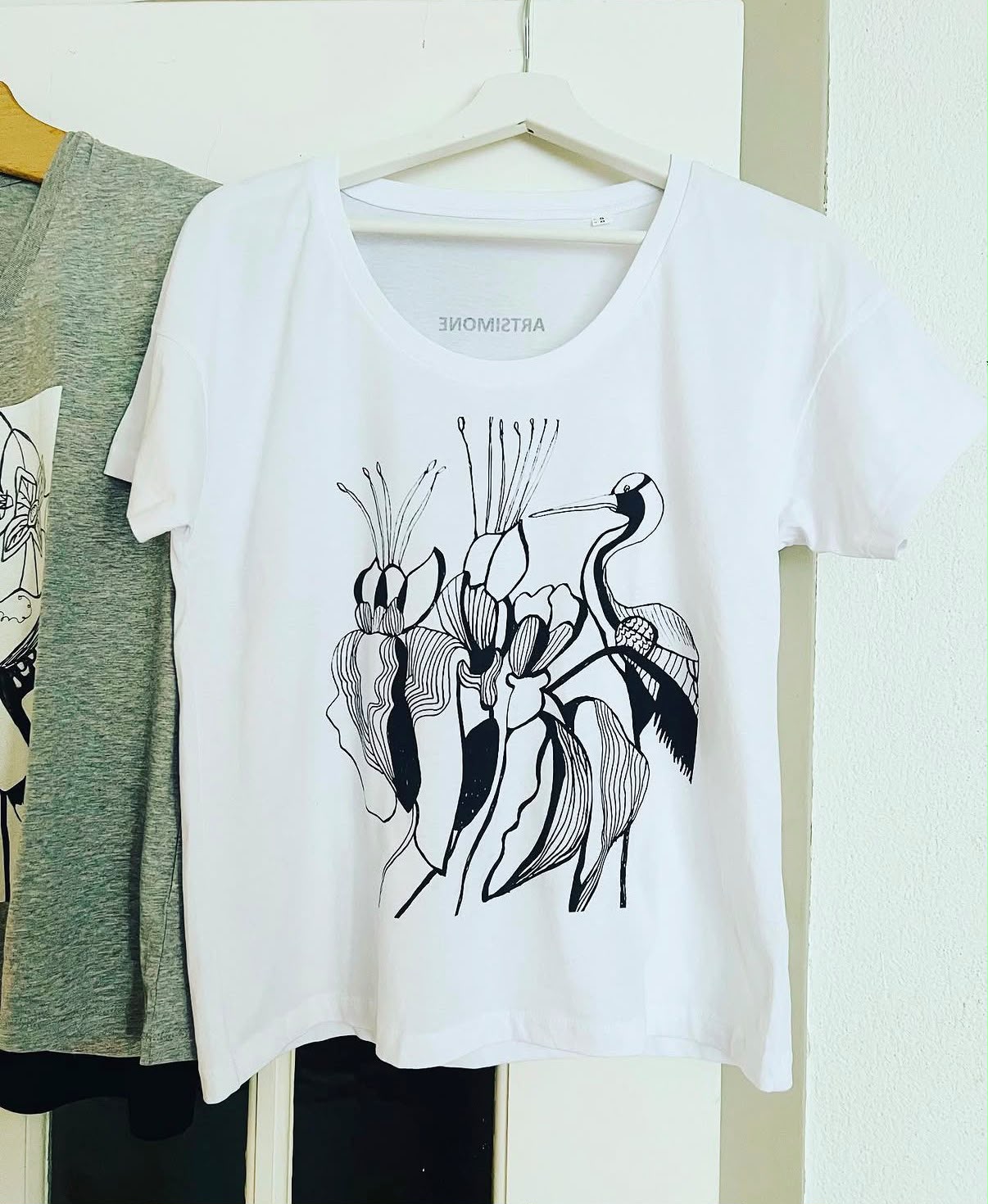 Weißes T-Shirt auf einem Bügel, bedruckt mit einer schwarzen Linienzeichnung aus Blüten mit langen Staubgefäßen und einem Kranich